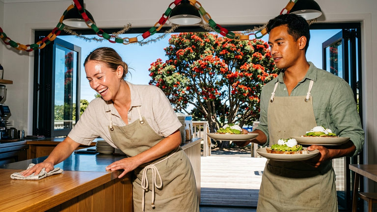 Two café workers in linen aprons preparing avocado toast, with festive paper chains and a pohutukawa tree visible through the window.