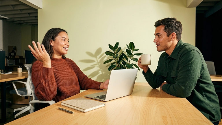 Two people chatting over a laptop and a coffee mug at a wooden table.