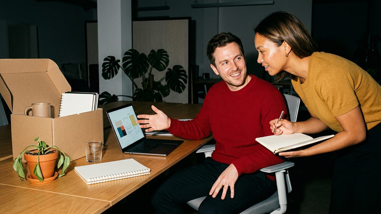Two people reviewing a laptop screen at a desk beside an open moving box packed with a mug, notebooks and a small plant.