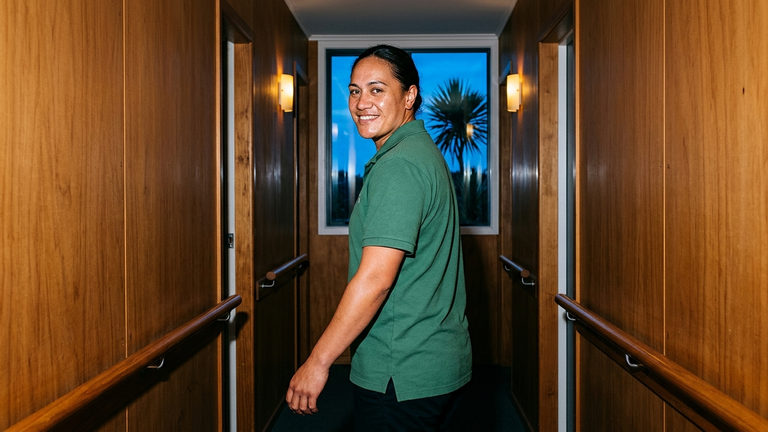 A support worker in a green polo shirt looking back over her shoulder in a wood-panelled care home hallway with handrails.