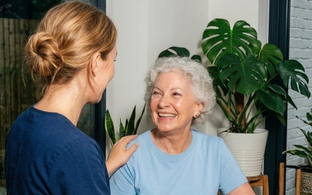 A support worker sitting at a kitchen table with an older woman, a hand on her shoulder, both of them laughing.