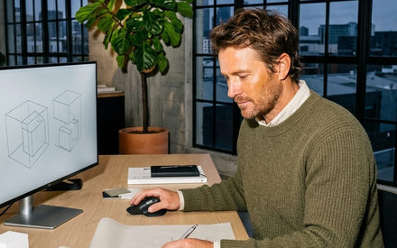 A man working at a desk in an office at dusk, drawing on a plan beside a monitor showing a design sketch.